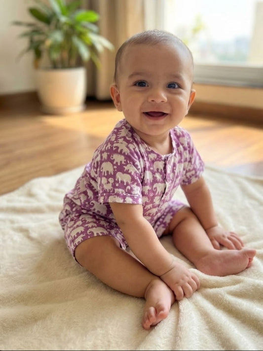 Baby sitting on a bed wearing a purple onesie with elephant patterns.