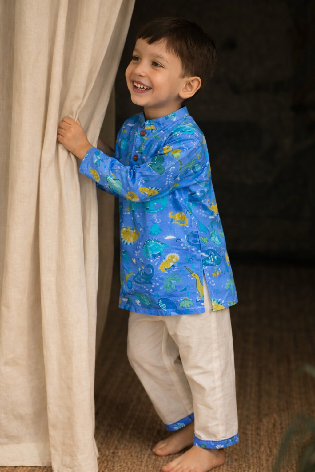 Child wearing a blue shirt with animal prints and beige pants peeking from behind a curtain.