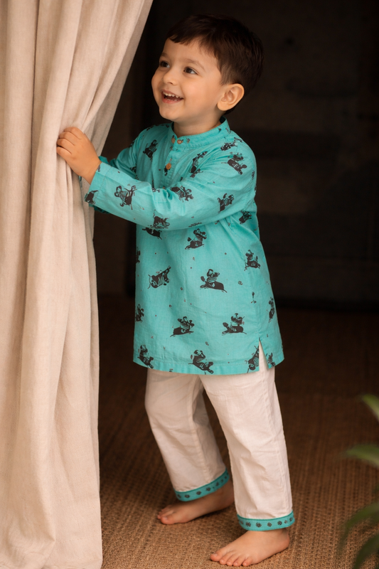 Child wearing a turquoise traditional outfit with white pants, standing behind a curtain.